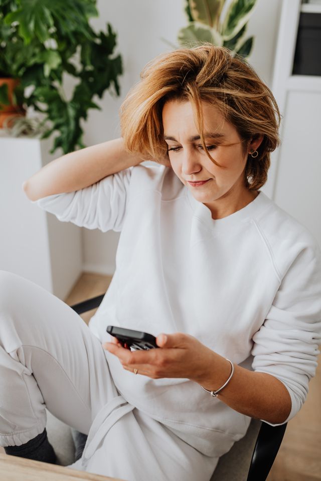 A relaxed woman in white attire using a smartphone indoors surrounded by plants.
